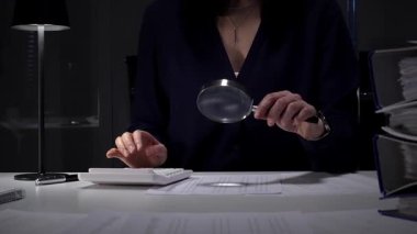 Vergi formlarını ve mali beyannameleri inceleyen kadın, hesap makinesi ve büyüteç kullanarak rakamları ve denetim kayıtlarını kontrol ediyor. İş dünyasında denetim ve vergi teması