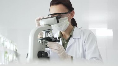 Mikroskop kaydırağını inceleyen koruyucu ekipmanlı kadın bilim adamı steril laboratuvar ortamında detaylı bilimsel araştırmalar yapıyor. Tıp, sağlık ve bilim konsepti