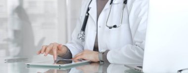 Beyaz önlüklü ve steteskoplu doktor dijital tablet kullanıyor, çevrimiçi danışmanlık yapıyor, hasta verilerini yenilikçi teknolojiyle yönetiyor. İlaç ve sağlık hizmetleri