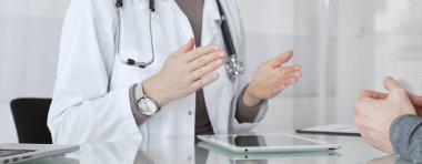 Klinik muayenesi sırasında bir hastaya tıbbi bilgileri açıklarken el hareketi yapan beyaz önlüklü kadın bir doktor. İlaç ve sağlık hizmetleri