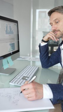 Takım elbiseli işadamı masa başına odaklanmış, klavyeye yazarken aynı zamanda kalemle not alıyor, modern ofis ortamında birden fazla iş yapıyor. İş adamları konsepti