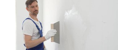 Koruyucu eldivenler ve mavi inşaat tulumları giyen erkek inşaat işçisi, tadilat projesi sırasında profesyonel alçı levha bantlı bıçak kullanarak duvarlara sorunsuzca alçı yayıyor. Portre