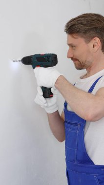 Mavi tulumlu, güvenlik gözlüklü ve iş eldivenli bir inşaat işçisi, kablosuz tornavida kullanarak duvara vida sokuyor ve dikkatli bir ev tadilatı yapıyor. Dikey portre