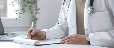 Masada çalışan, reçete yazan, beyaz laboratuvar önlüğü giyen ve klinik çalışma alanında steteskop kullanan bir erkek doktor. Kapatın. İlaç ve sağlık hizmetleri kavramı.