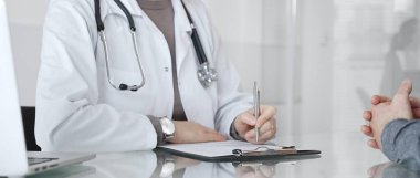 Klinikteki hasta muayenesi sırasında panoya not alan beyaz önlüklü doktor sağlık, teşhis ve bakım kararlarına odaklanmış. İlaç ve sağlık hizmetleri