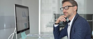 Profesyonel iş adamı gözlük takıyor ve spor ceket giyiyor, çağdaş bir ofis ortamında çalışıyor, bir masaüstü bilgisayarında görevler yapıyor. İş adamları konsepti