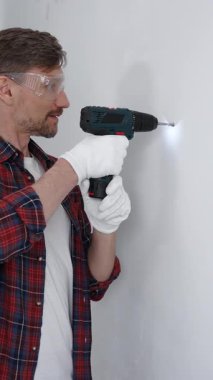 Kırmızı tişört ve beyaz eldiven giyen orta yaşlı erkek işçi, ev yenileme çalışmaları sırasında kablosuz matkapla duvarda delik açıyor. Dikey görünüm.