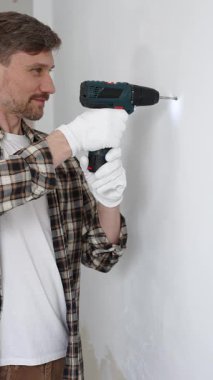 İşaretli gömlek ve beyaz eldiven giyen orta yaşlı bir adam, ev yenileme çalışmaları sırasında kablosuz matkapla duvarda delik açıyor. Dikey görünüm.