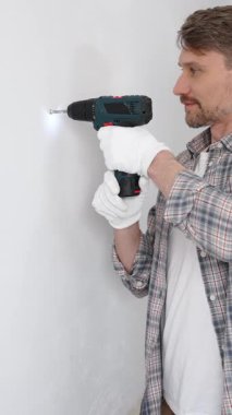Gri gömlekli ve beyaz eldivenli orta yaşlı işçi, ev yenileme çalışmaları sırasında kablosuz matkapla duvarda delik açıyor. Dikey görünüm.