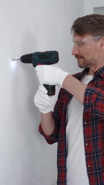 Kırmızı tişört ve beyaz eldiven giyen orta yaşlı erkek işçi, ev yenileme çalışmaları sırasında kablosuz matkapla duvarda delik açıyor. Dikey görünüm.