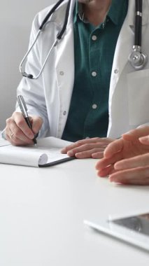 Yeşil gömlekli bir doktor, parlak ve modern bir klinikte muayene sırasında hastanın sağlık sorunlarını dinliyor ve reçete yazıyor. İlaç ve sağlık hizmetleri kavramı