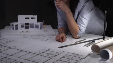 İnşaat planları üzerinde çalışan kadın mimar ve bir masanın üzerinde mimari tasarım ve geliştirme üzerine odaklanan bir model ev..