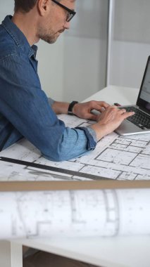 Mimar, yeni projeler için ayrıntılı planlama, inşaat ve tasarım mühendisliğini sembolize eden, kat planlarını gösteren bir cetvel ve dizüstü bilgisayar kullanarak beyaz bir masanın üzerine bina planları çiziyor.