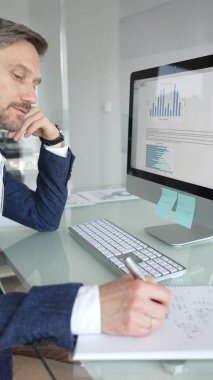 Takım elbiseli işadamı masa başına odaklanmış, klavyeye yazarken aynı zamanda kalemle not alıyor, modern ofis ortamında birden fazla iş yapıyor. İş adamları konsepti