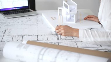 İnşaat planları ve çağdaş bir yapı modeli üzerinde çalışan kadın mimar, masasında yenilikçi tasarımlar ve kentsel kalkınma stratejileri kavramlaştırıyor.