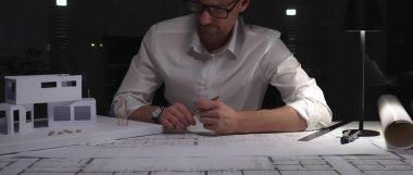 Adam mimar, gece karanlık bir ofiste planlar ve ölçekli bir model üzerinde çalışırken yeni bir mimari bina yaratıyor. İş ve mimarlık kavramı