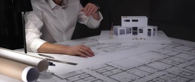 Mimar planlar üzerinde yoğunlaşır, titizlikle yeni bir ev projesi için tasarımlar hazırlar, fiziksel bir model üzerinde çalışır. İş ve mimarlık kavramı