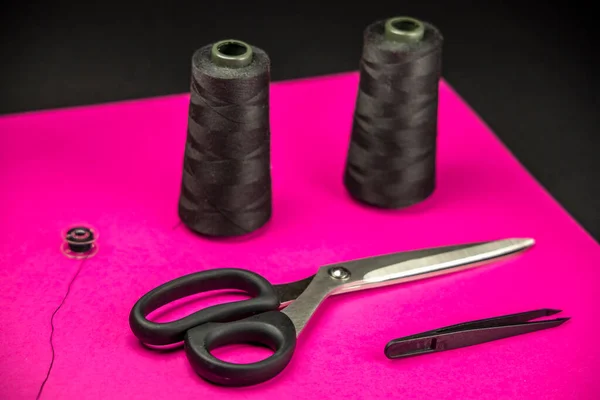 Black tailoring accessories as scissors, thread, tweezers and bobbin on pink background