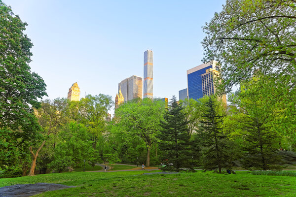 Green trees and Midtown Manhattan skyline viewed from Central Park South, in New York, USA. Tourists in the distance.