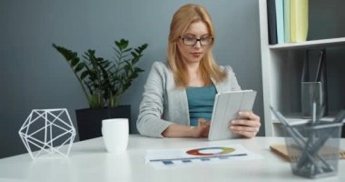 Genç, zeki, güzel, gözlüklü, masada oturan, elinde tabletle çalışan sarışın kadın..