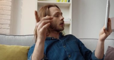 Kotlu tişörtlü, yakışıklı, beyaz bir adam video görüşmesinde elinde tabletle evde oturuyor..