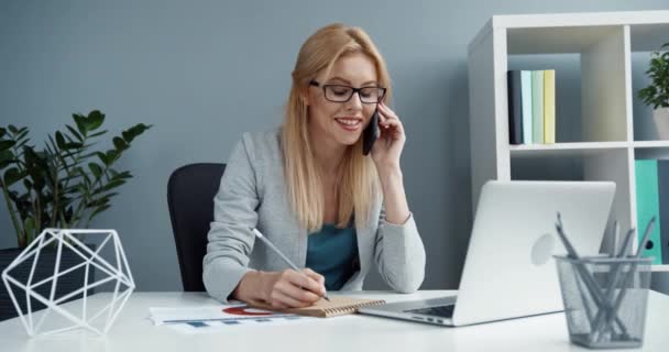 Mladý krásný blondýnka žena s brýlemi sedí v kanceláři v práci mluvit na mobilní telefon a psaní poznámek v notebooku. — Stock video