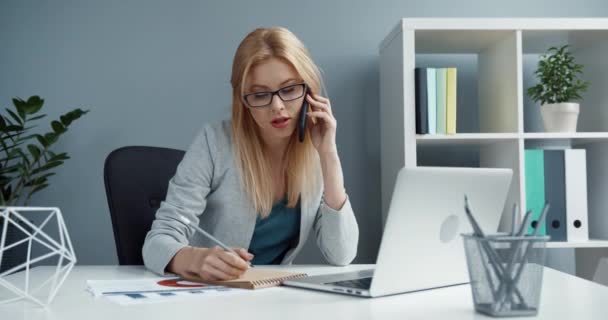 Mladá krásná blondýnka s brýlemi sedí v kanceláři na pracovišti a mluví na mobilu s klientem. Vzdálená konzultace manažera. — Stock video