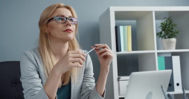 Jeune belle femme blonde intelligente avec des lunettes assis dans le bureau derrière le lieu de travail regardant attentivement dans la distance et tordant crayon dans les mains. — Video