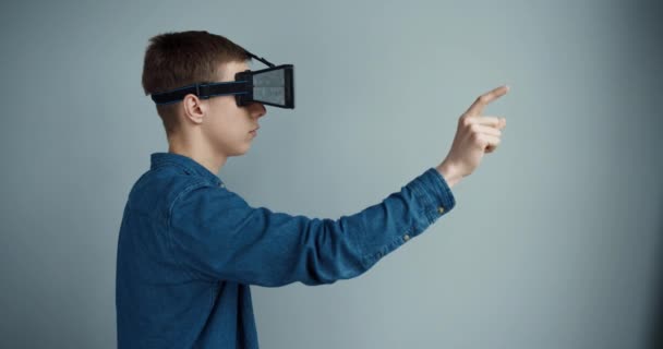 Молодой парень в джинсовой рубашке стоит на заднем плане стены в шлеме виртуальной реальности и использует жесты для работы с виртуальной реальности. Вид сбоку. — стоковое видео