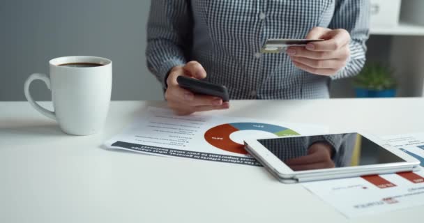 Close-up van vrouwelijke handen op bureaubladachtergrond met kopje koffie en tablet met bankkaart in handen en invoeren van gegevens op smartphone. — Stockvideo