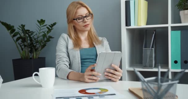 Jonge succesvolle blonde zakenvrouw met bril zitten in het kantoor aan een bureau met kopje koffie en werken holding tablet kijken uit het raam. — Stockvideo