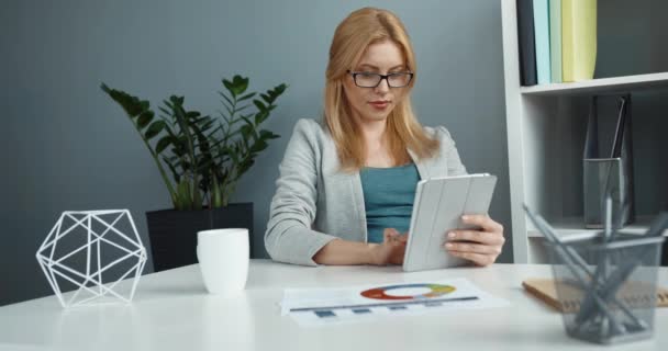 Jeune belle femme blonde intelligente avec des lunettes assis au bureau avec une tasse de café et travaille tenant la tablette. — Video