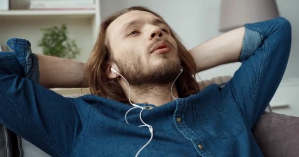 Mladý pohledný běloch s dlouhými vlasy v džínové košili odpočívá na gauči a poslouchá hudbu na sluchátkách. — Stock video