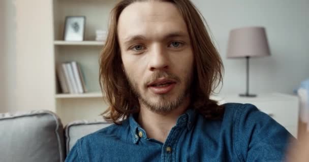 Young handsome caucasian guy with long hair in denim shirt is talking on video call while sitting at home on the couch. Camera view. — Stock Video
