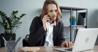 Uzun saçlı, genç, şık bir ofis çalışanı işte oturuyor cep telefonuyla konuşuyor ve dizüstü bilgisayar kullanıyor..