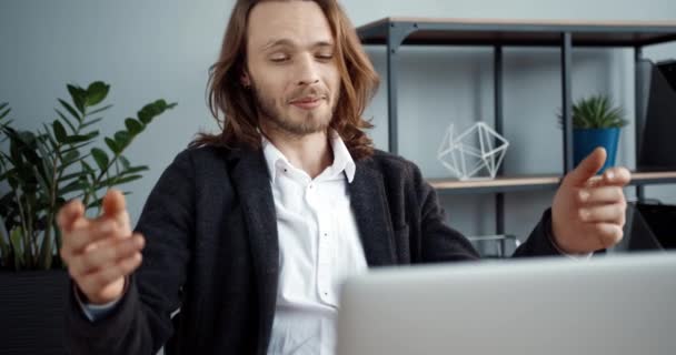 Молодой успешный кавказский бизнесмен с длинными волосами общается через видео на своем ноутбуке, находясь в офисе. — стоковое видео