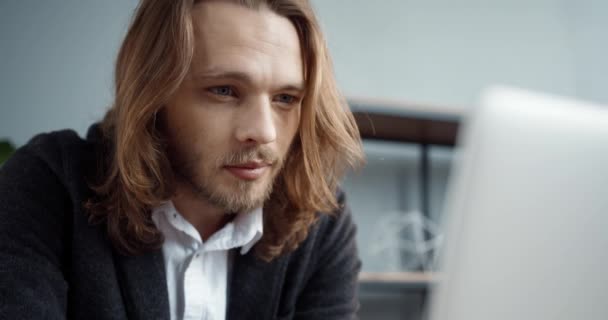 Nahaufnahme eines jungen erfolgreichen, gut aussehenden Geschäftsmannes mit langen Haaren, der im Büro sitzt und intensiv in seinem Laptop arbeitet. — Stockvideo