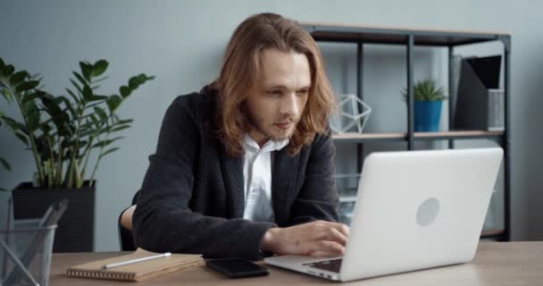Intelligente giovane uomo d'affari di successo con i capelli lunghi seduto in ufficio e lavorando intensamente nel suo computer portatile su un nuovo progetto. — Video Stock