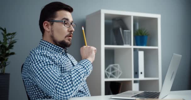 Jovem funcionário de escritório caucasiano com óculos se senta no local de trabalho e pensa em resolver o problema, e escreve a ideia no caderno. — Vídeo de Stock