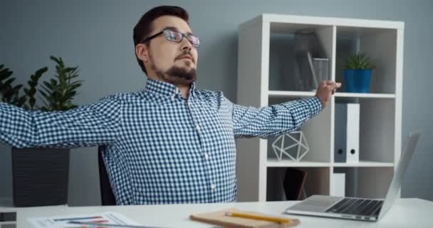 Inteligentny młody pracownik biurowy w koszuli w kratę i okularach rozciąga się siedząc przy biurku i odpoczywając po wykonanej pracy. — Wideo stockowe