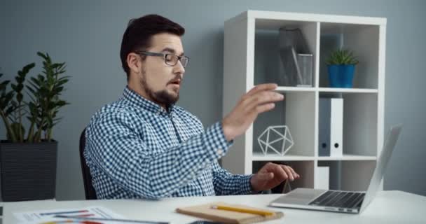 穿着格子呢衬衫和眼镜的快乐的办公室职员坐在办公桌旁的笔记本电脑上完成了他的工作，然后伸出手来. — 图库视频影像