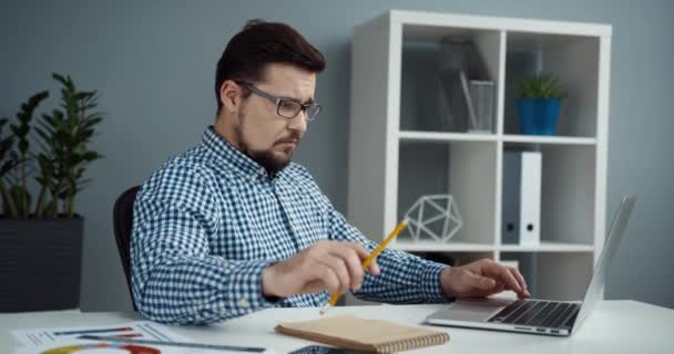 격자무늬 셔츠와 안경을 착용하고 일하는 성공적 인 사무직 근로자는 책상에 앉아 노트북 컴퓨터를 사용하고 노트북으로 노트를 작성 한다. — 비디오