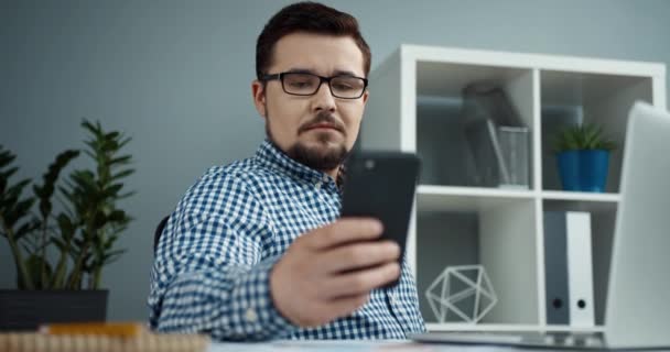 Giovane direttore d'ufficio in occhiali con barba si siede in ufficio moderno e risponde al messaggio sul suo smartphone. — Video Stock