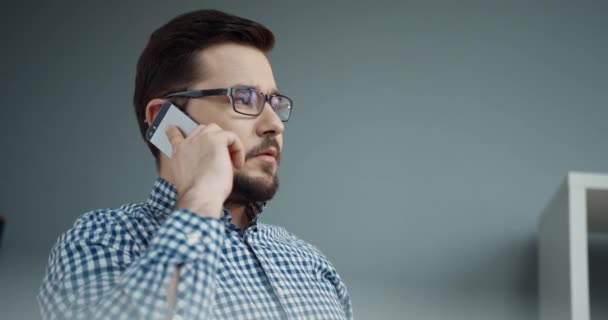 オフィスに座って、携帯電話で話している間に注文を与える眼鏡を持つ白人ビジネスマンの閉鎖. — ストック動画