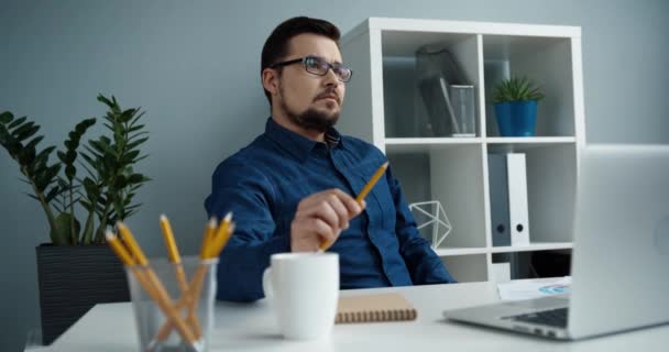 Inteligente trabalhador de escritório caucasiano com óculos senta-se no local de trabalho e pensa em resolver o problema torcendo lápis em sua mão. — Vídeo de Stock