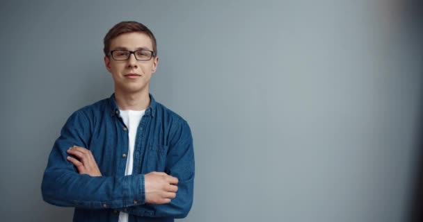 Porträtt av ung smart stilig kille i jeansskjorta och glasögon tittar på kameran med armarna korsade och ler. — Stockvideo