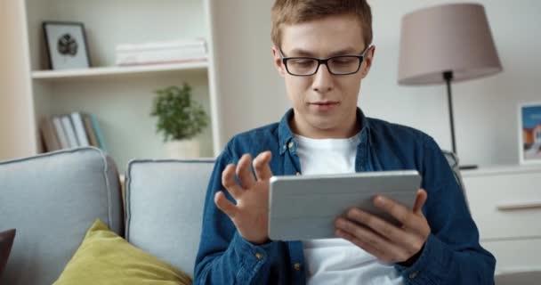 Jovem bonitão de camisa jeans jogando videogames usando tablet enquanto sentado em casa no sofá. — Vídeo de Stock