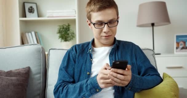 Jonge knappe jongen in denim shirt is chatten met vrienden op sociale netwerken met behulp van smartphone terwijl thuis zitten op de bank. — Stockvideo