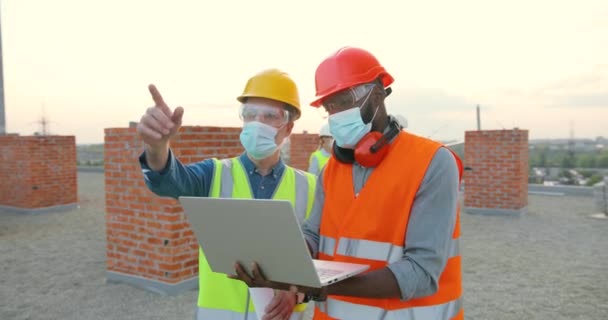 身穿医疗面罩、护目镜和手提电脑站在室外建筑工地的混血年轻人。讨论工作的多族裔男性建筑工人、工人和建筑工人. — 图库视频影像