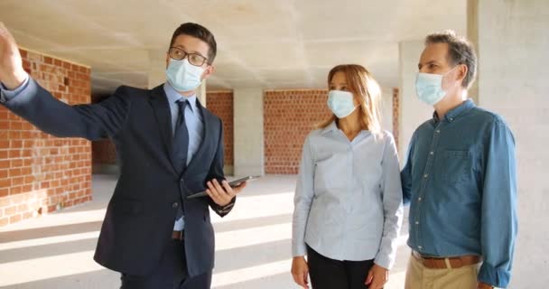 Kaukasischer männlicher Immobilienmakler zeigt verheirateten Kunden in medizinischen Masken eine Wohnung in der Bauphase und spricht Frau und Mann, die eine neue Wohnung auf der Baustelle kaufen. Coronavirus — Stockvideo
